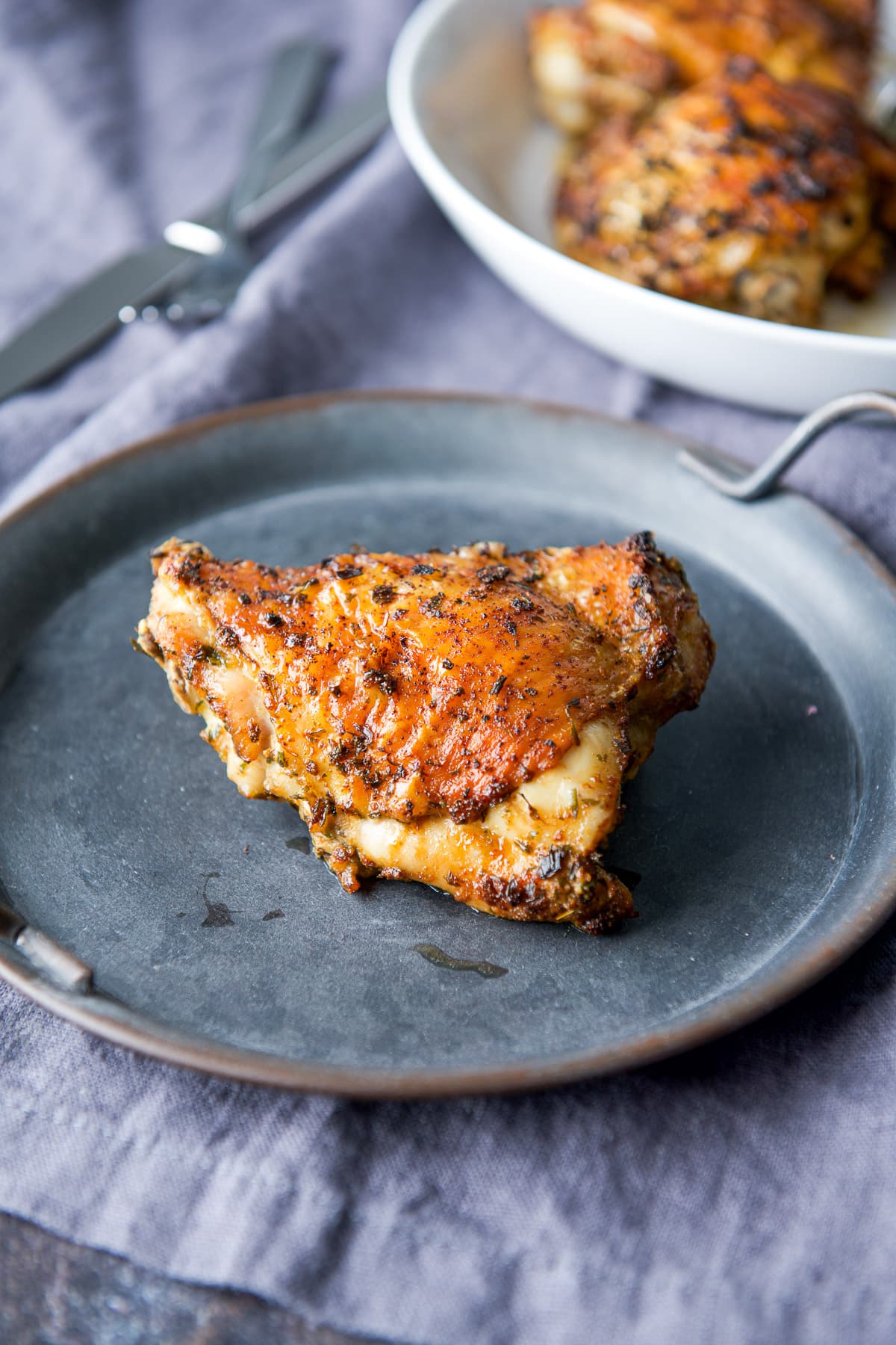 A cooked chicken thigh with crispy skin on a metal plate with more thighs in the background