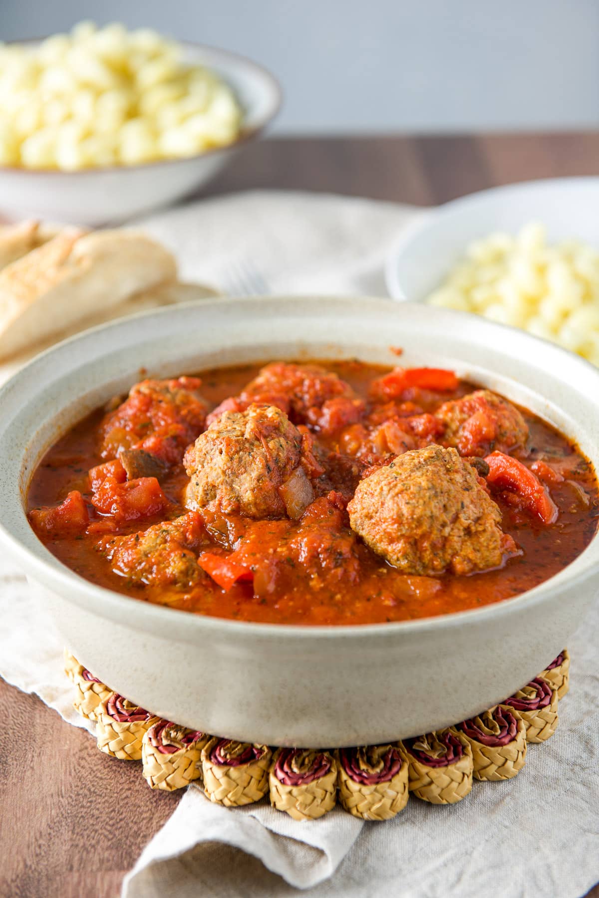 A grey bowl with sauce and meatballs with pasta and bread behind