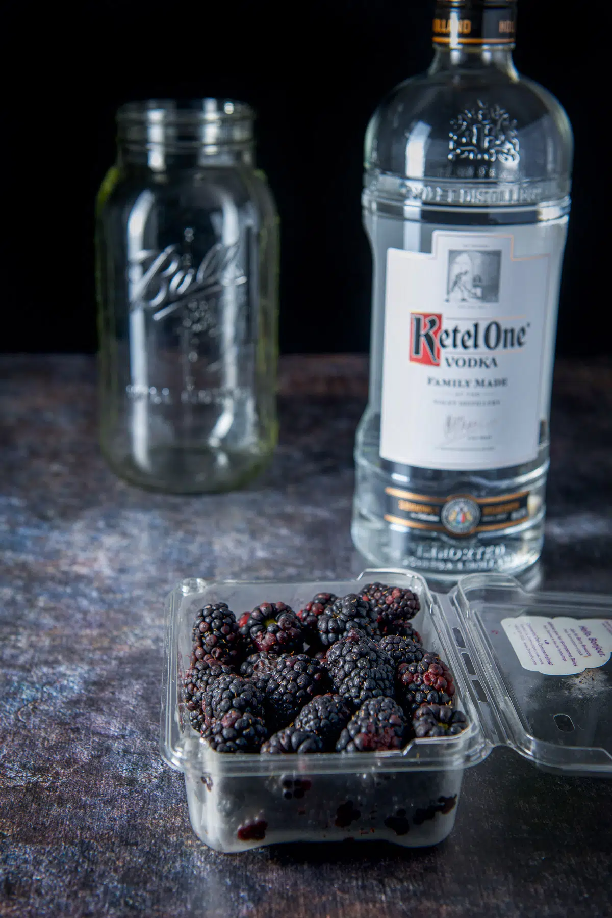 A plastic container of blackberries, bottle of vodka and a jar