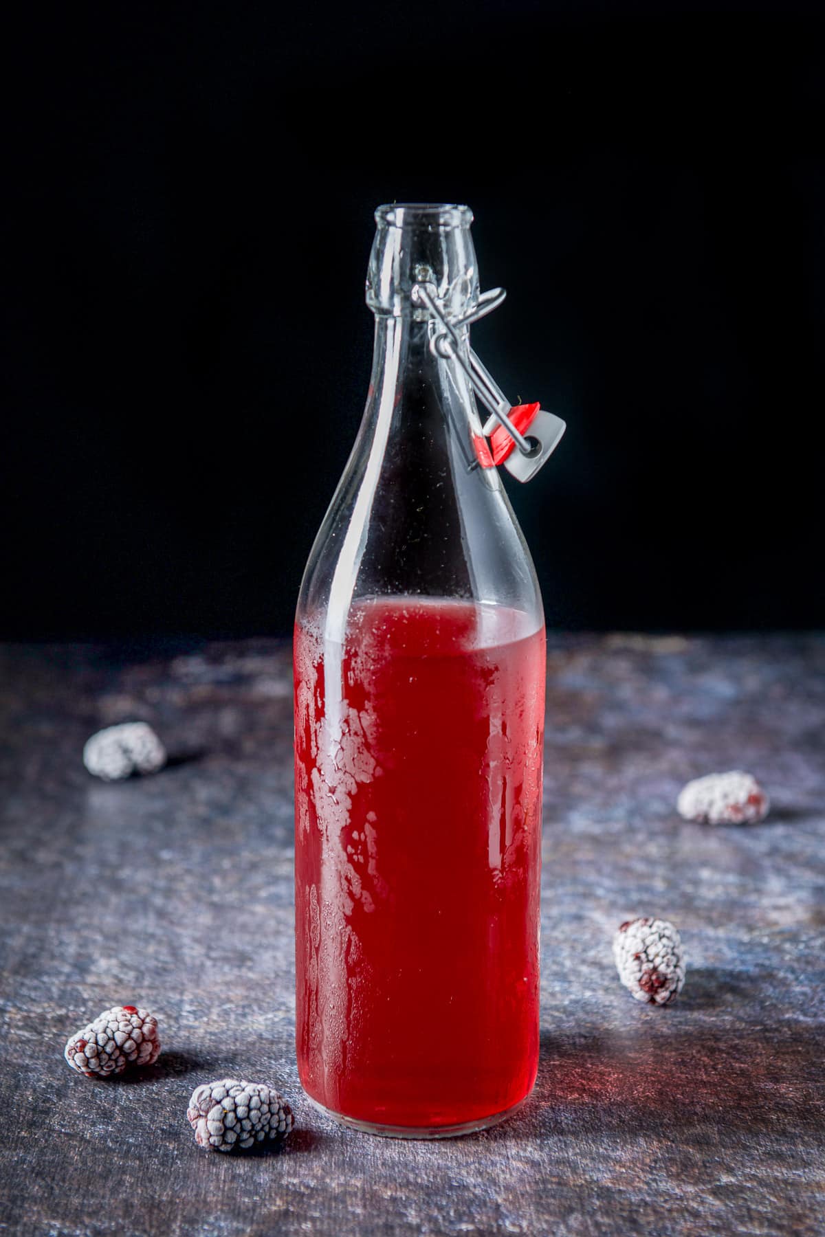 A frosty bottle with red infused vodka with 5 blackberries on a table