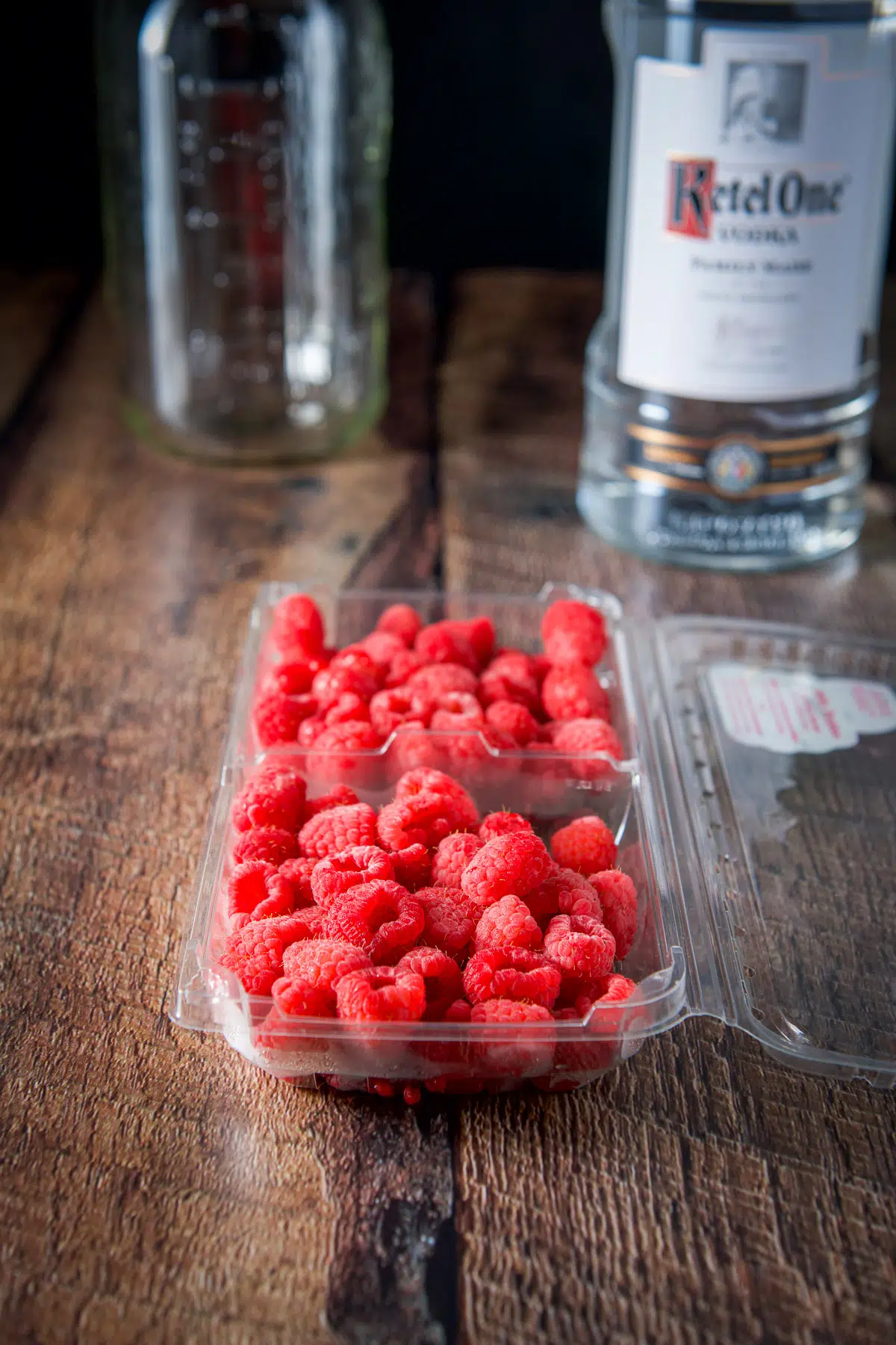 Raspberries in a plastic container, a vodka bottle and jar in the background