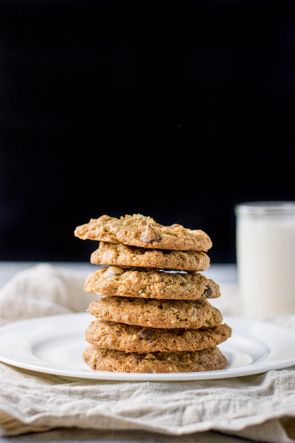 A white plate with a stack of cookies with a glass of milk in the back