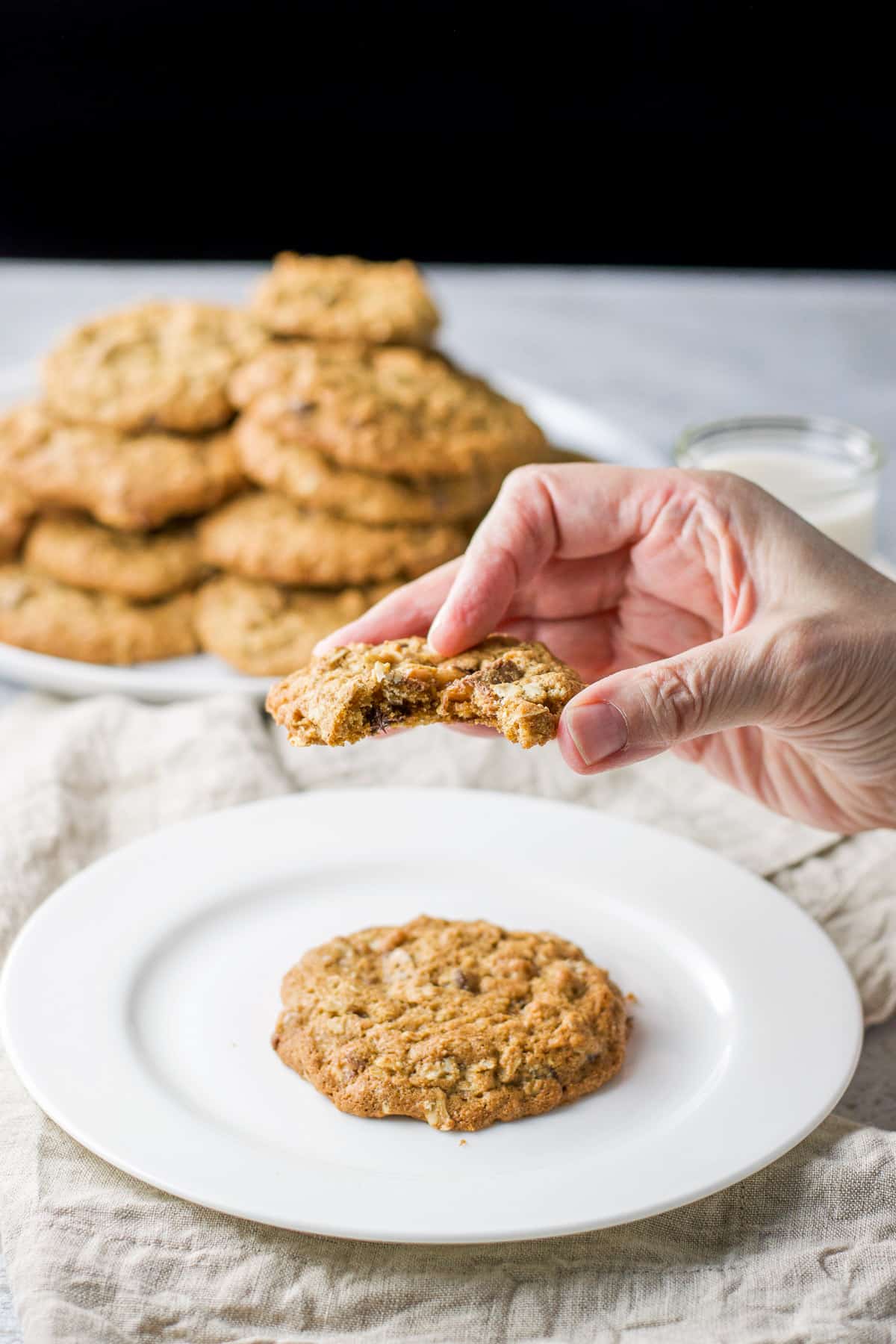 A hand holding a cookie with a bite taken out of it. There is a pile of cookies on a plate in the back