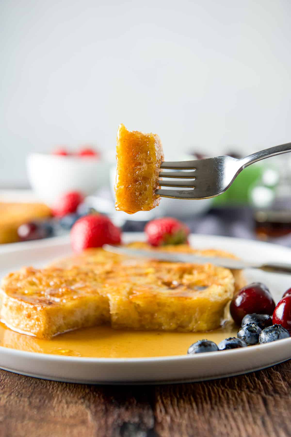 A fork holding a piece of egg toast over a plate with slices and maple syrup