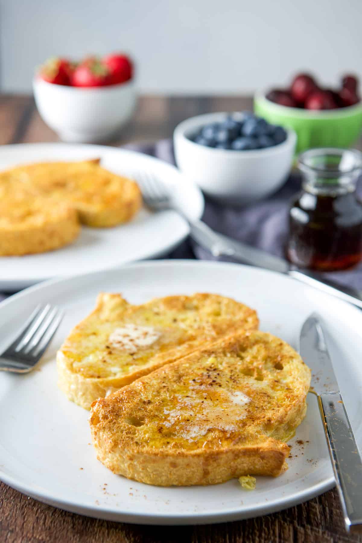 Plates with the French toast with melted butter and cinnamon sprinkled; fruit and maple syrup in back