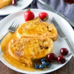 French toast smothered with maple syrup on a white plate with cherries, strawberries and blueberries