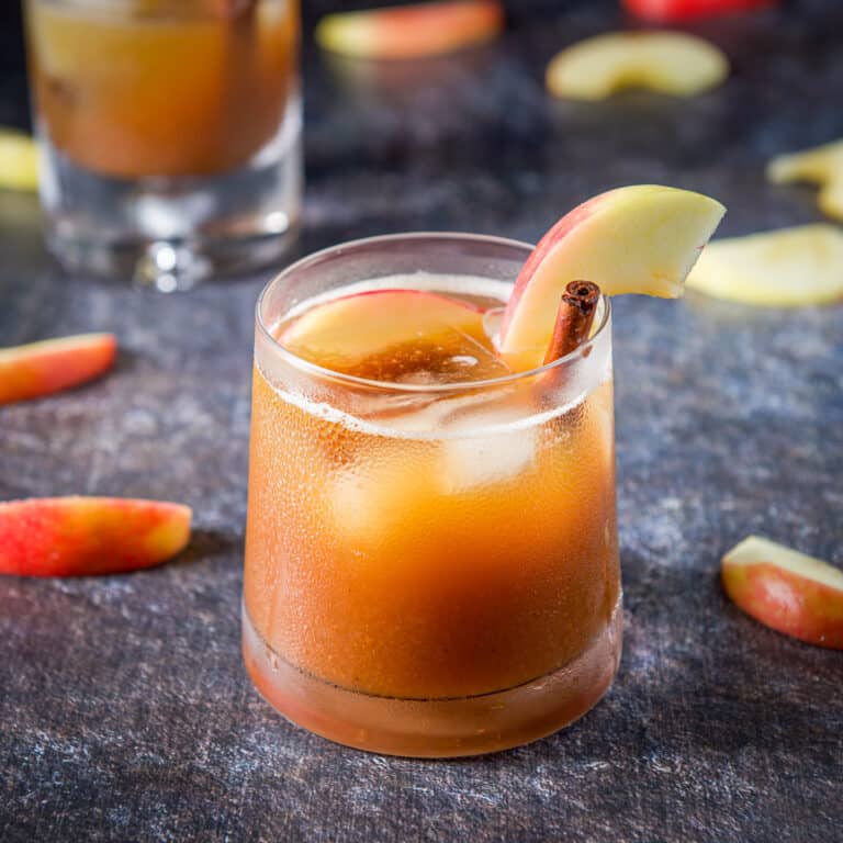 A sloped glass filled with the apple cocktail with an apple slice and cinnamon stick as garnish