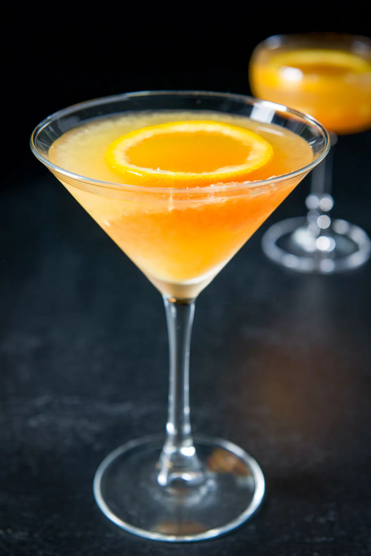 Martini glass with the orange cocktail in it with a coupe glass in the back