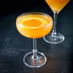 The orange cocktail in a coupe glass with an orange rind as garnish - square