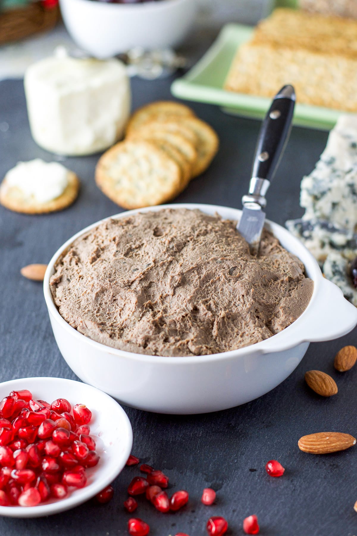 Close up of the white bowl with the liver pate and some pomegranate seeds cheeses and crackers in the background