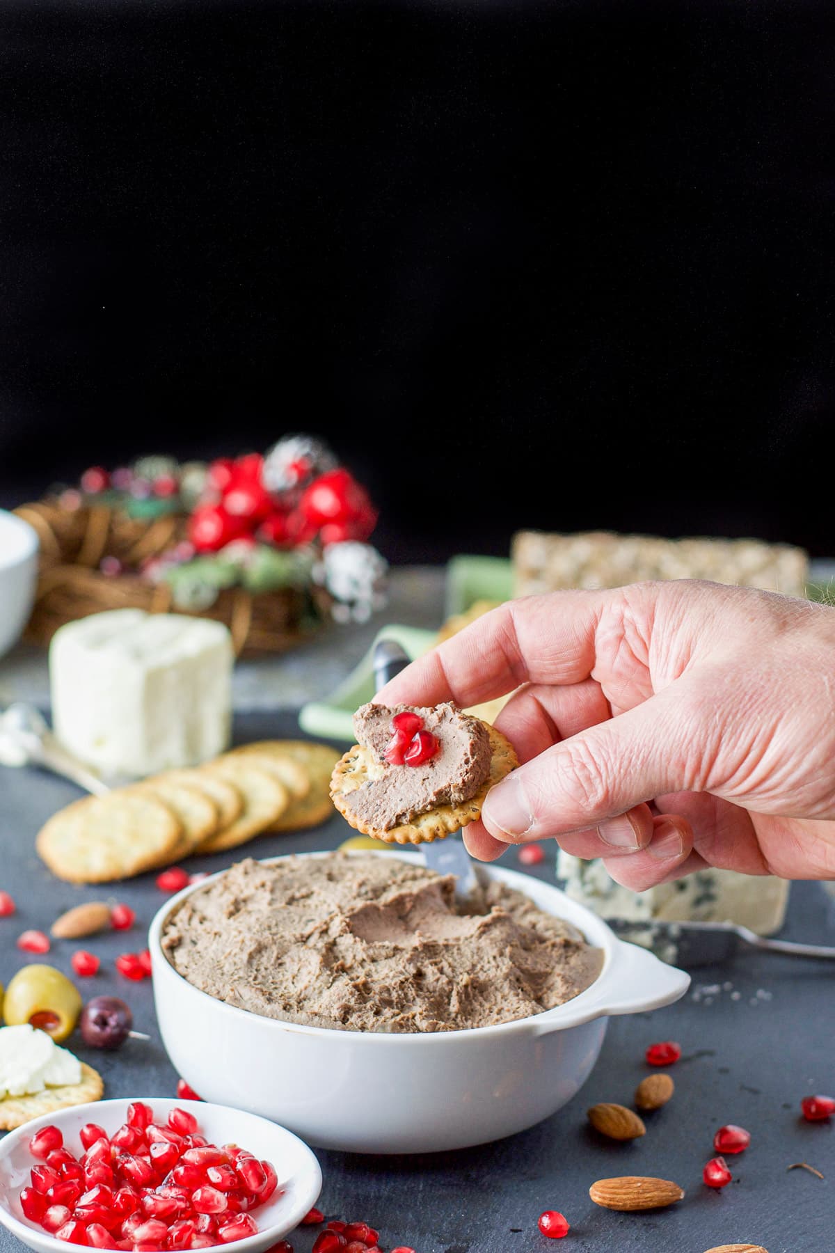 A male hand holding a cracker with pate on it over the bowl with lots of appetizers in the background