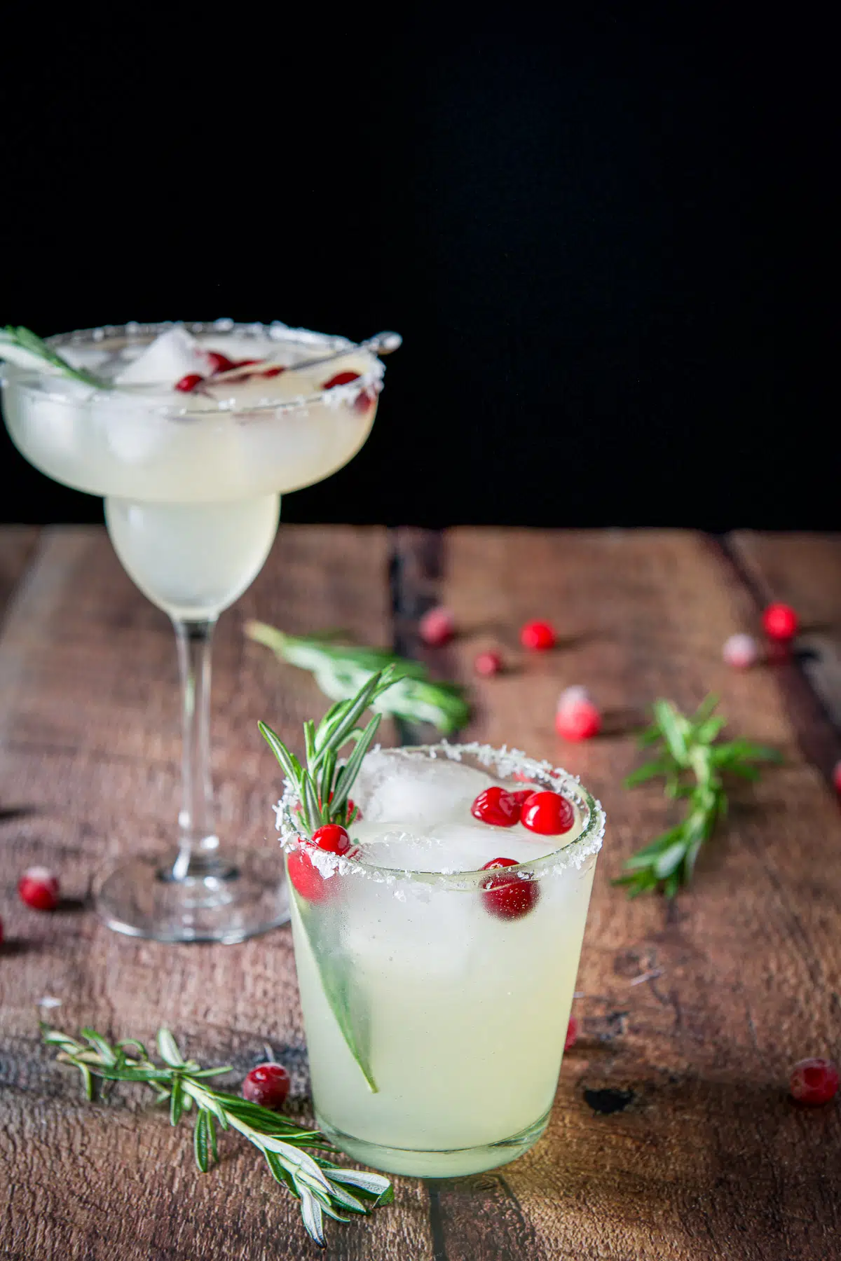 A short glass in front of the classic glass, both filled with the margarita. Cranberries and rosemary are the garnish