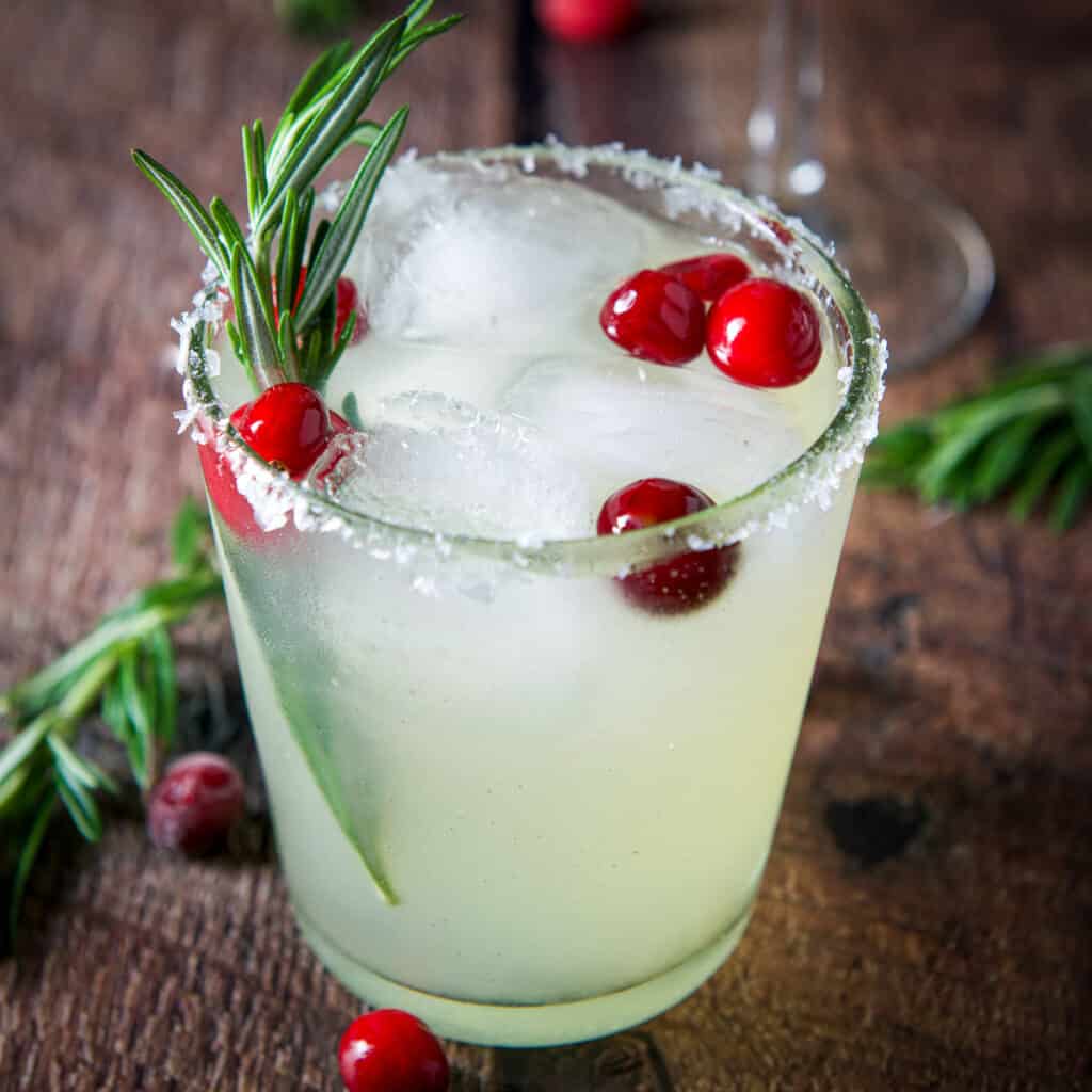 A double old fashioned glass with the margarita in it with cranberries and rosemary sprigs - square