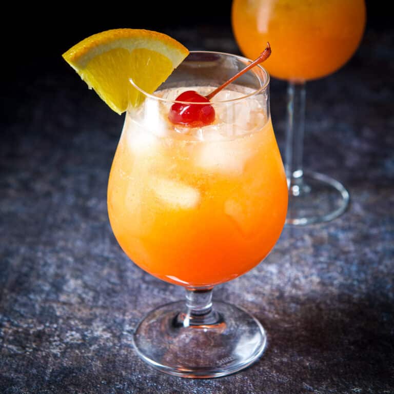 The colorful cocktail in a tulip glass with a cherry and orange slice on the rim - square