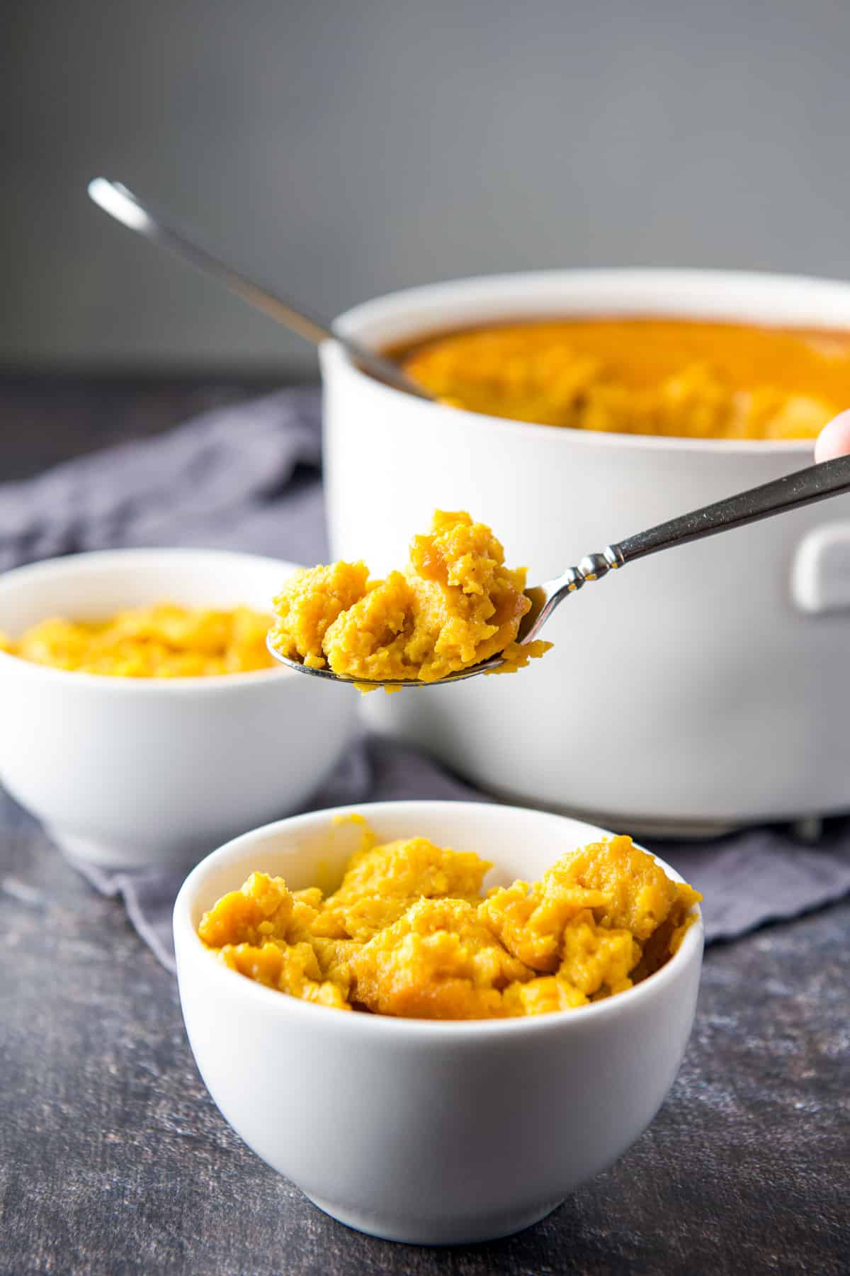 A spoon with some souffle on it, held over a small bowl with another bowl and a casserole dish in the back