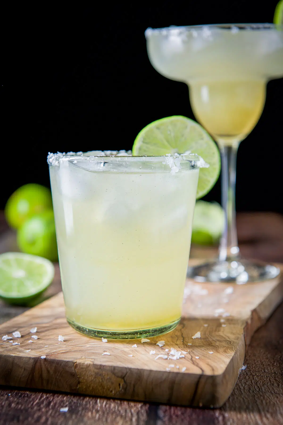 Vertical view of two margarita glasses filled with amber liquid and limes in the back