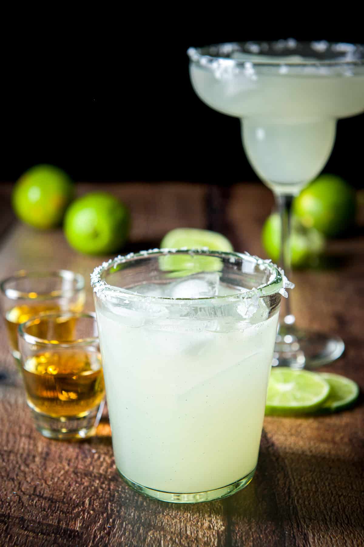 Two salted glasses with limes in the back filled with margaritas with shots of Grand Marnier off the side