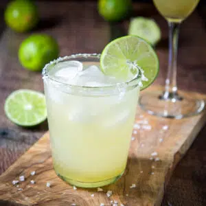 A wooden board with a glass with a margarita in it, limes in the back