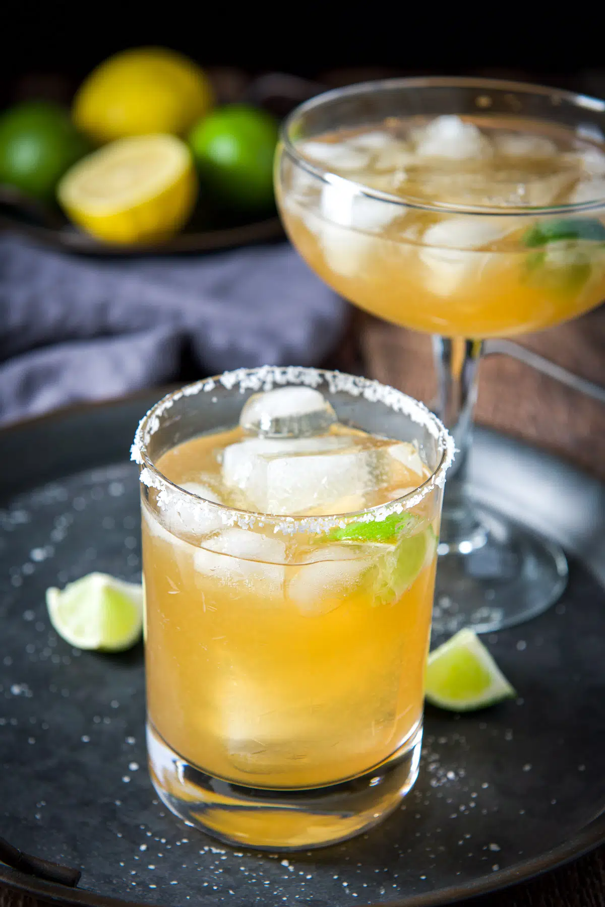 A salted glass in front of a bowl glass with margaritas in them with lemon and lime behind