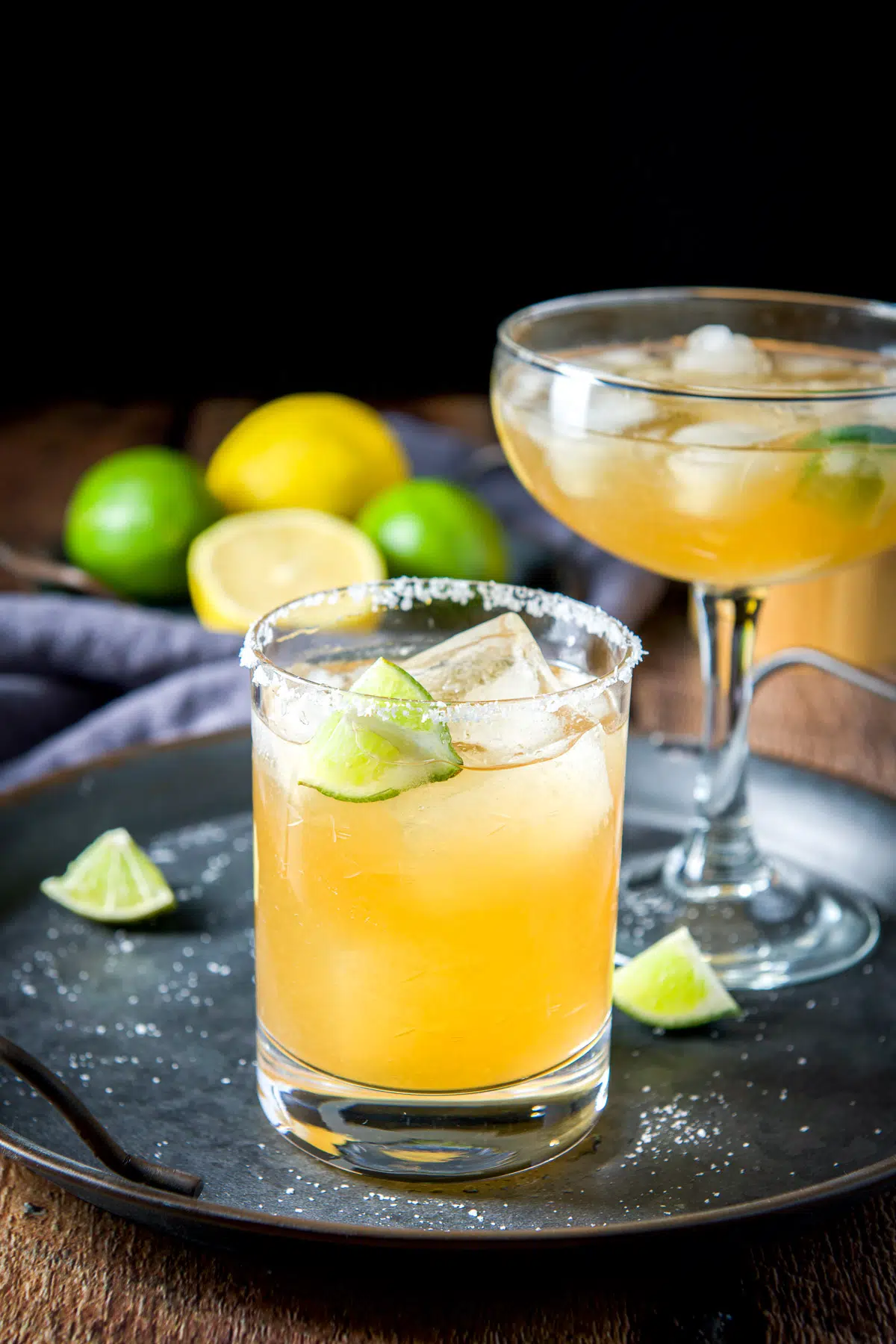 A metal tray with lime and salt strewn and two glasses with margaritas in them