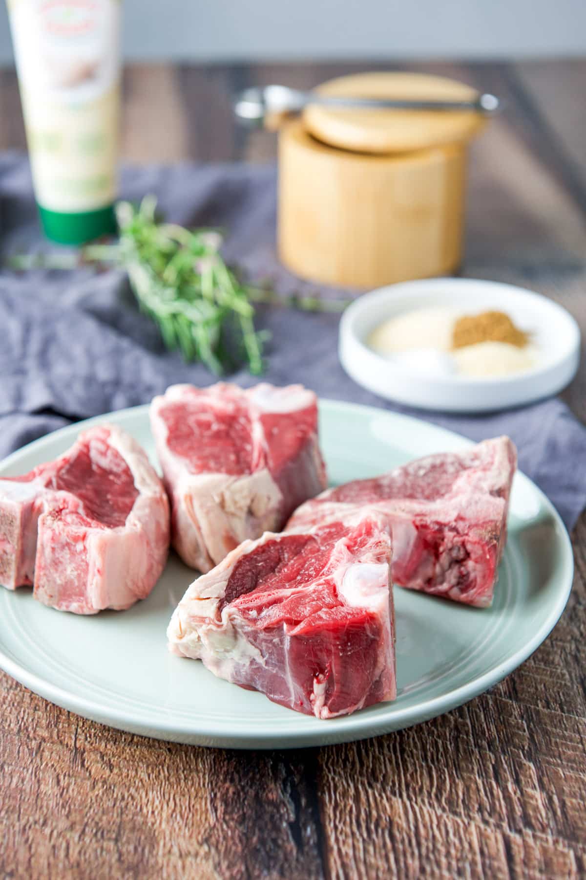 A small green plate with raw lamb chops, with fresh herbs, a plate of spices and salt in back
