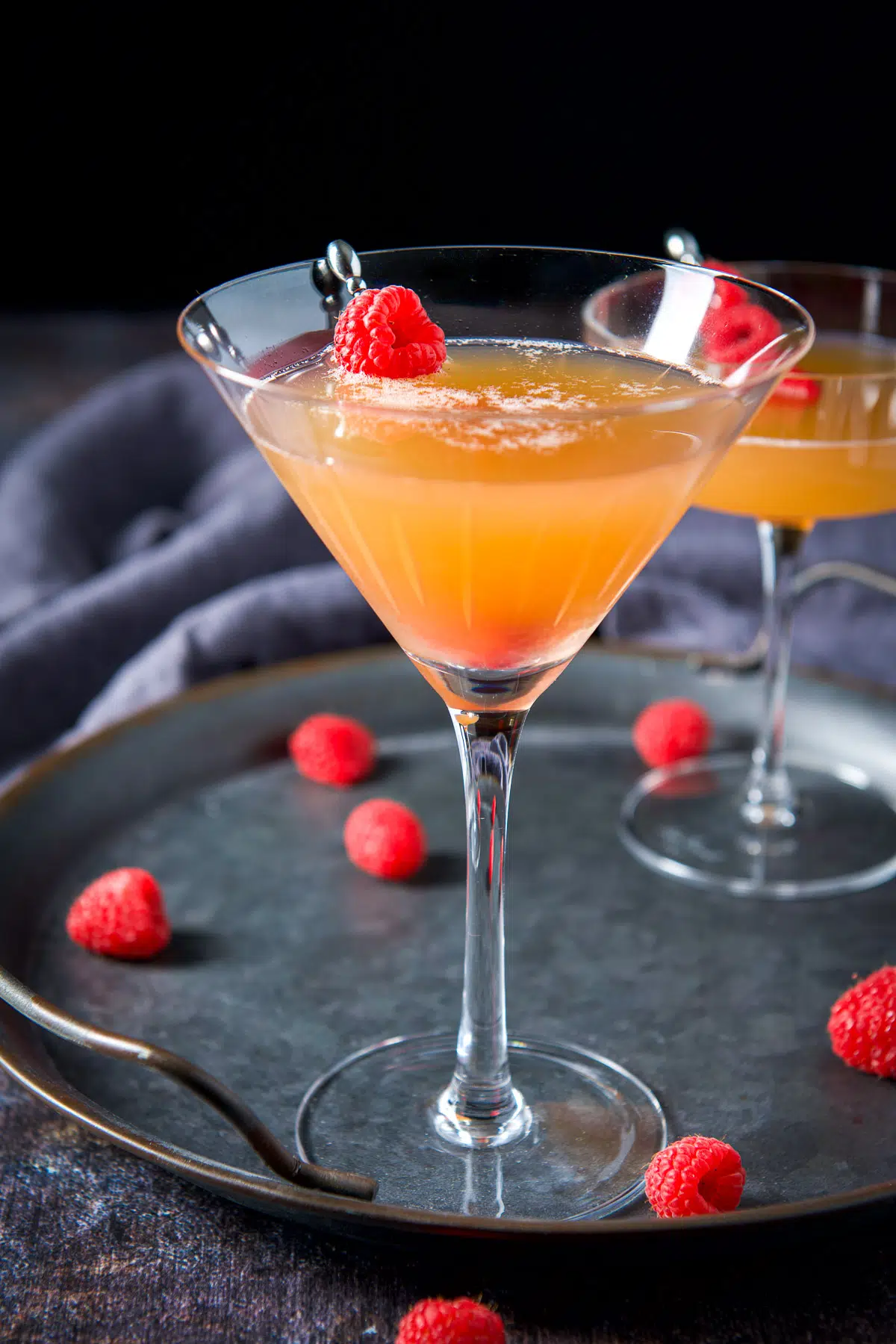 A metal tray with two cocktails on it with raspberries on the tray and in the drink