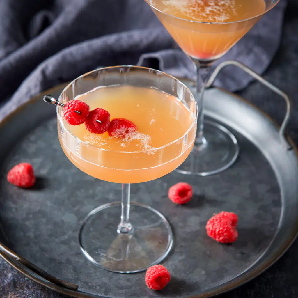 Square photo of two cocktail glasses filled with a amber martini with raspberries on the tray and in the drinks