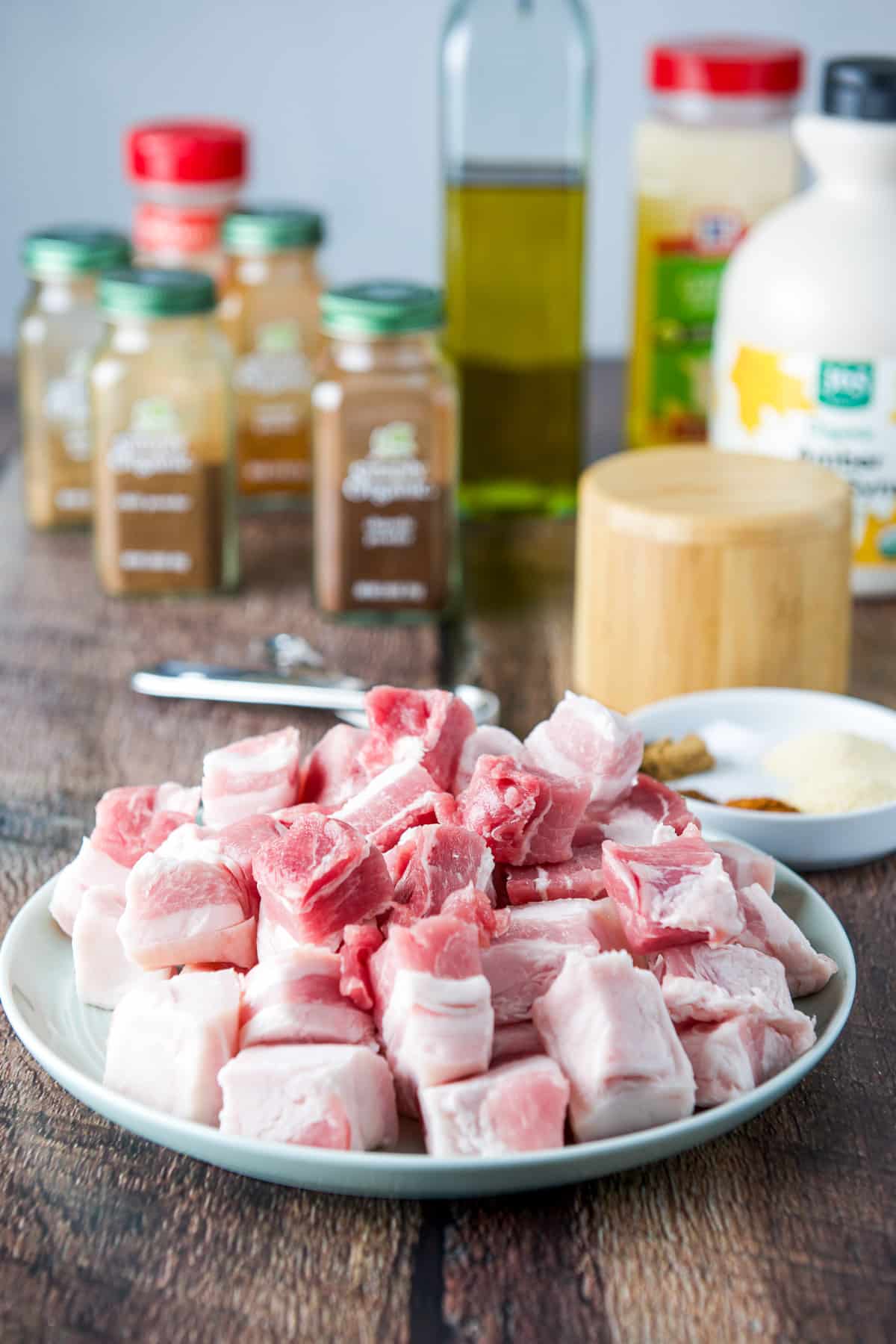A pile of raw pork bellies on a plate with herbs, spices, oil and maple syrup in the back