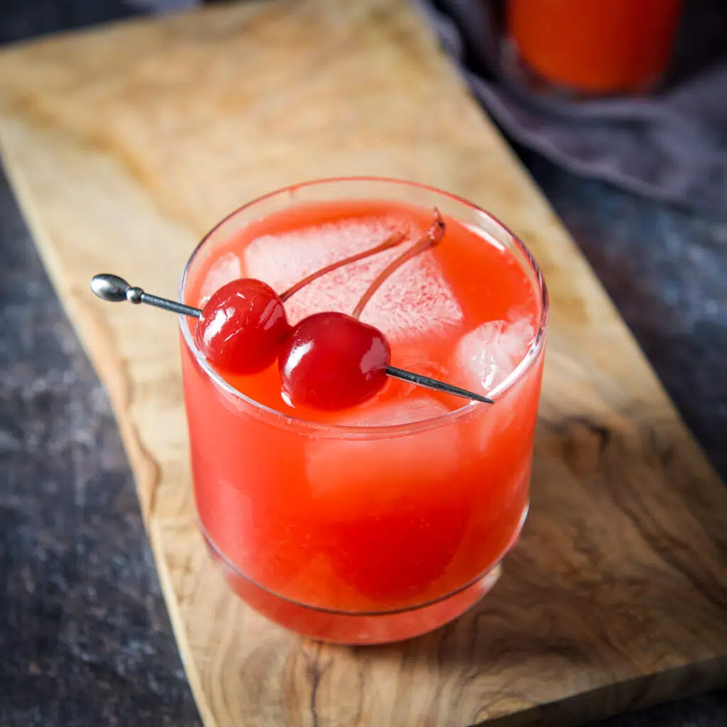 A glass with a red cocktail with two maraschino cherries on a pick - square