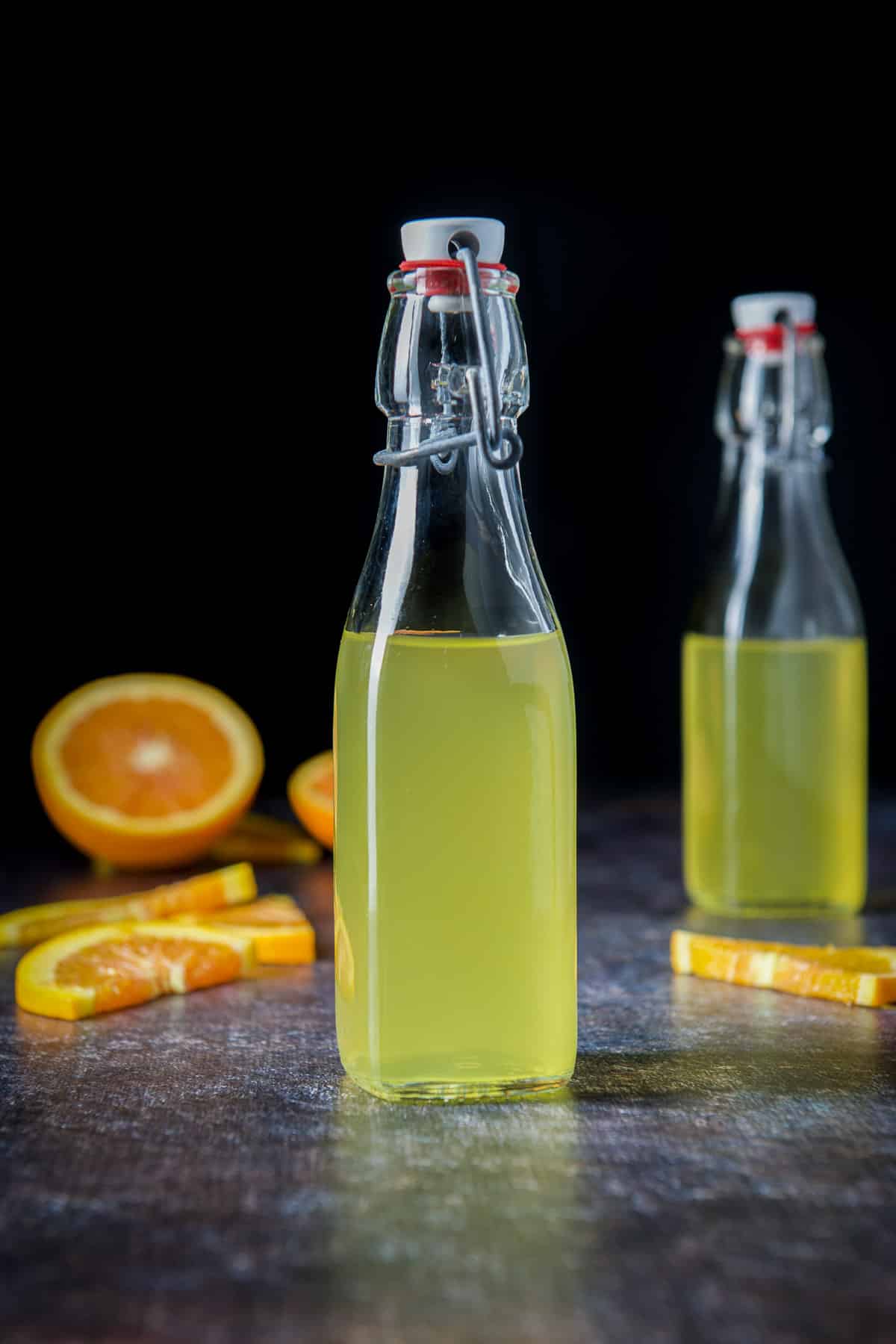 Two small glass bottles with vodka in them and sliced oranges in the back