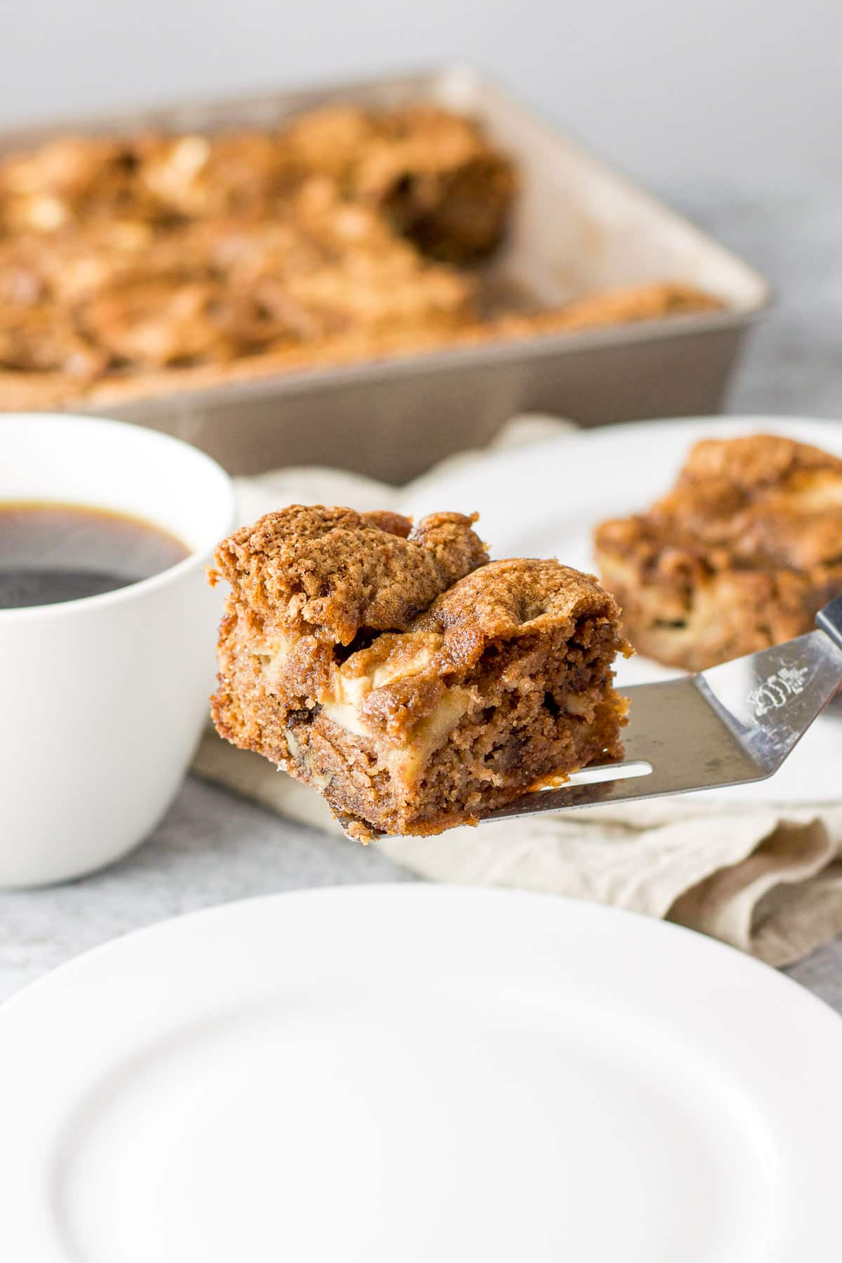 Piece of cake held over a white plate with coffee and the rest of the cake in back