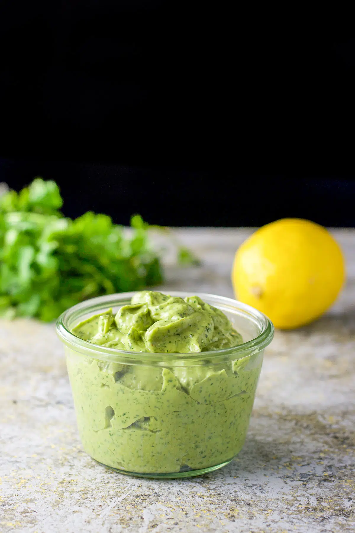 A glass jar with the green dip in it and cilantro and lemon in the back
