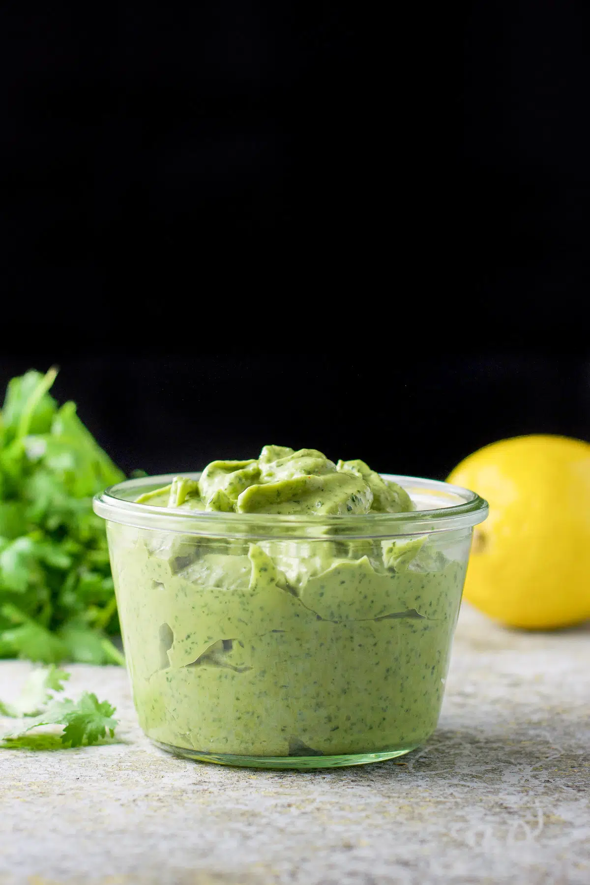 Vertical view of a jar of heaping avocado dip in it with lemon and cilantro in the back