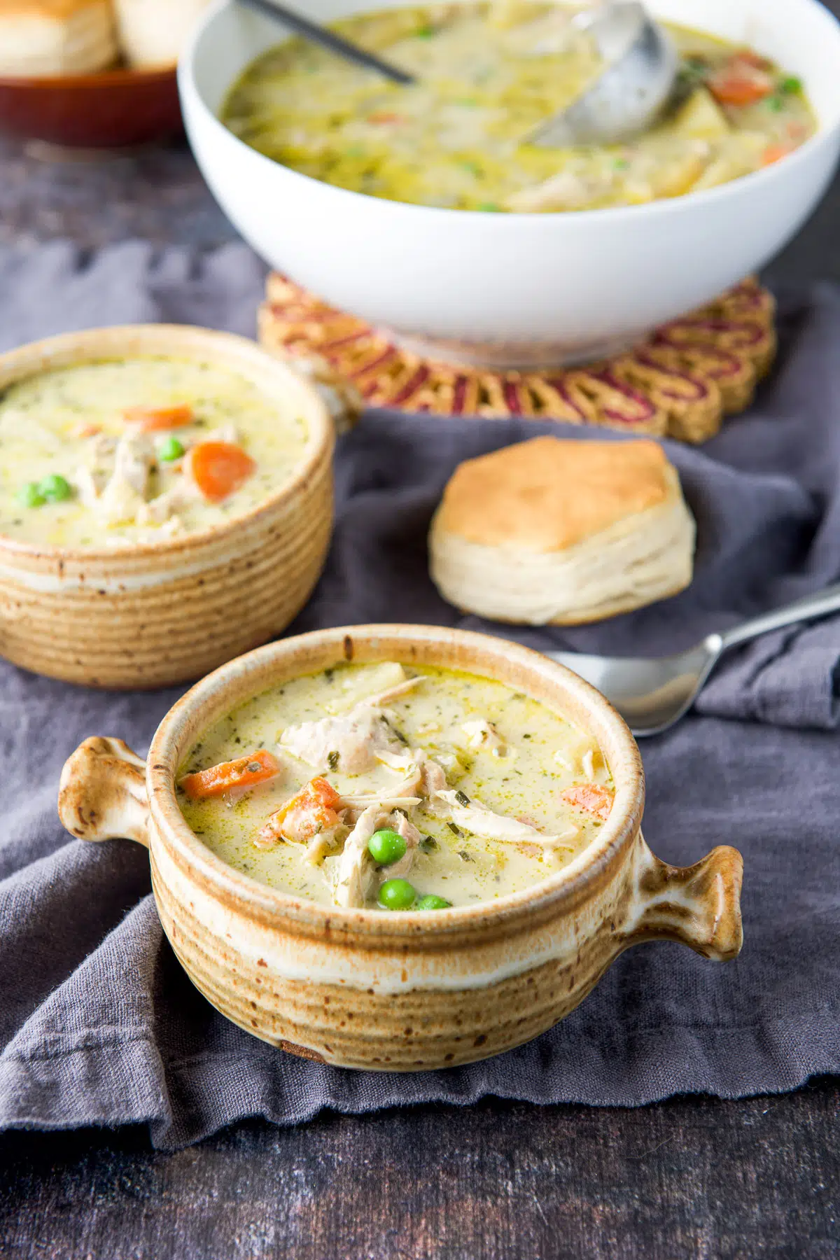 Two bowls of chicken soup in front of a spoon, biscuit and serving bowl