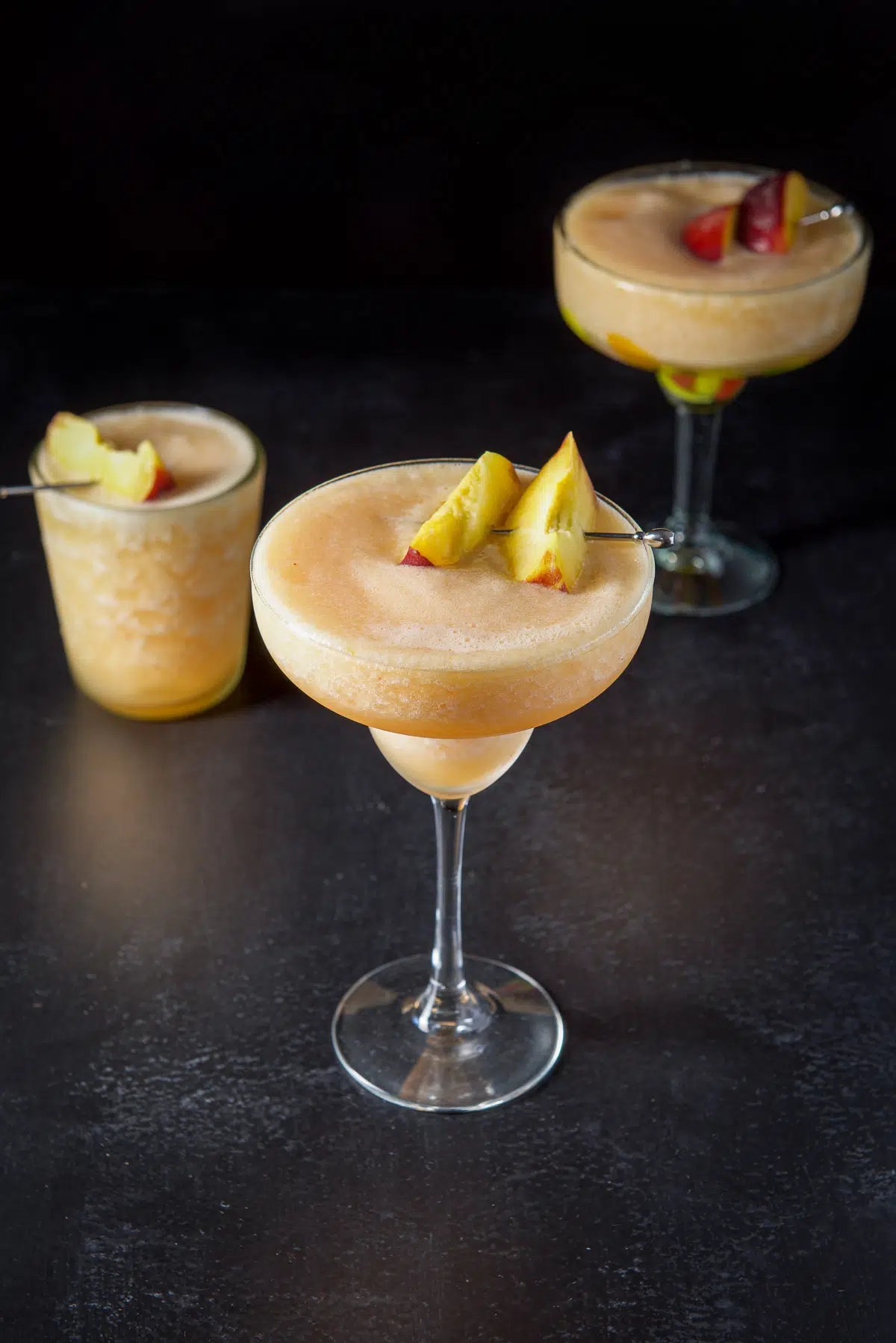 A classic margarita glass filled with the margarita and fresh peach slices with two other glasses in the back
