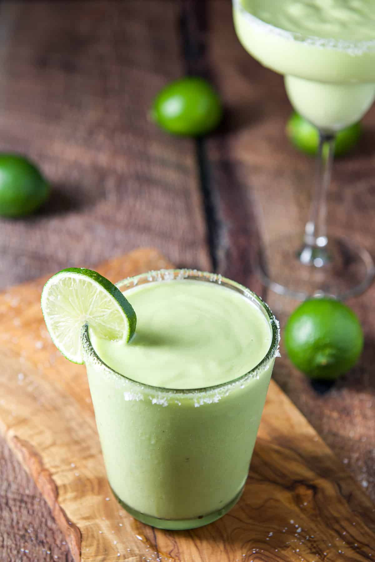 A short margarita glass with the green margarita on a board with salt on the table along with limes