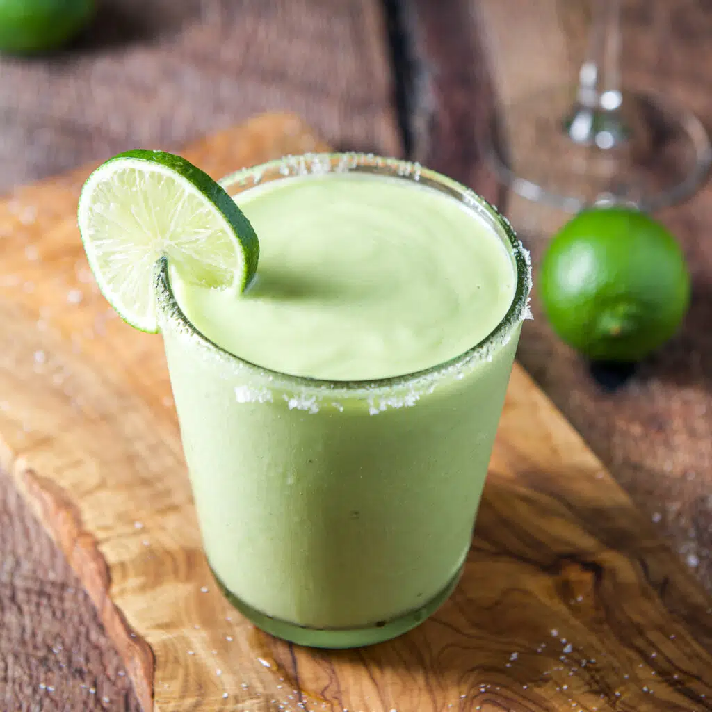 Square photo of a salted avocado margarita on a board with lime