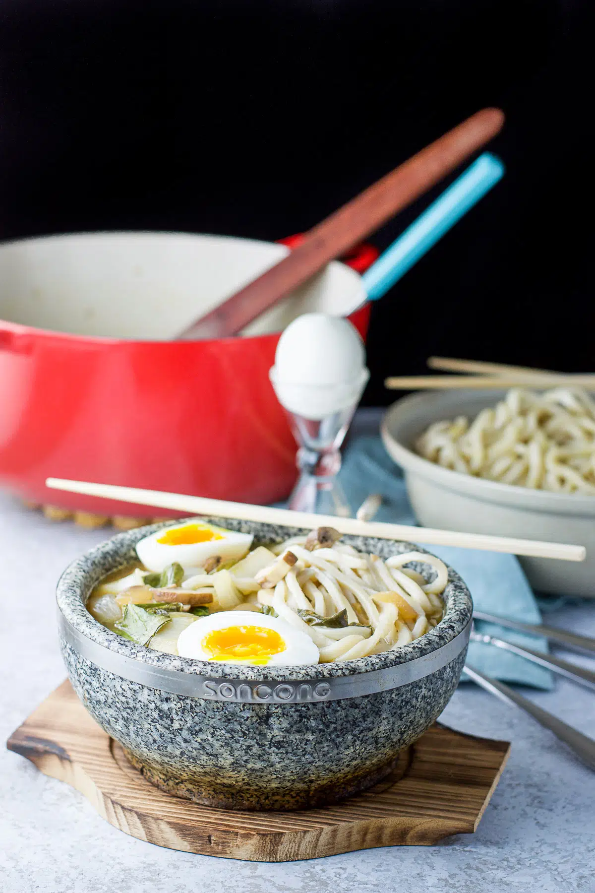 Stone bowl filled with noodle soup with eggs in it along with the pan in the back