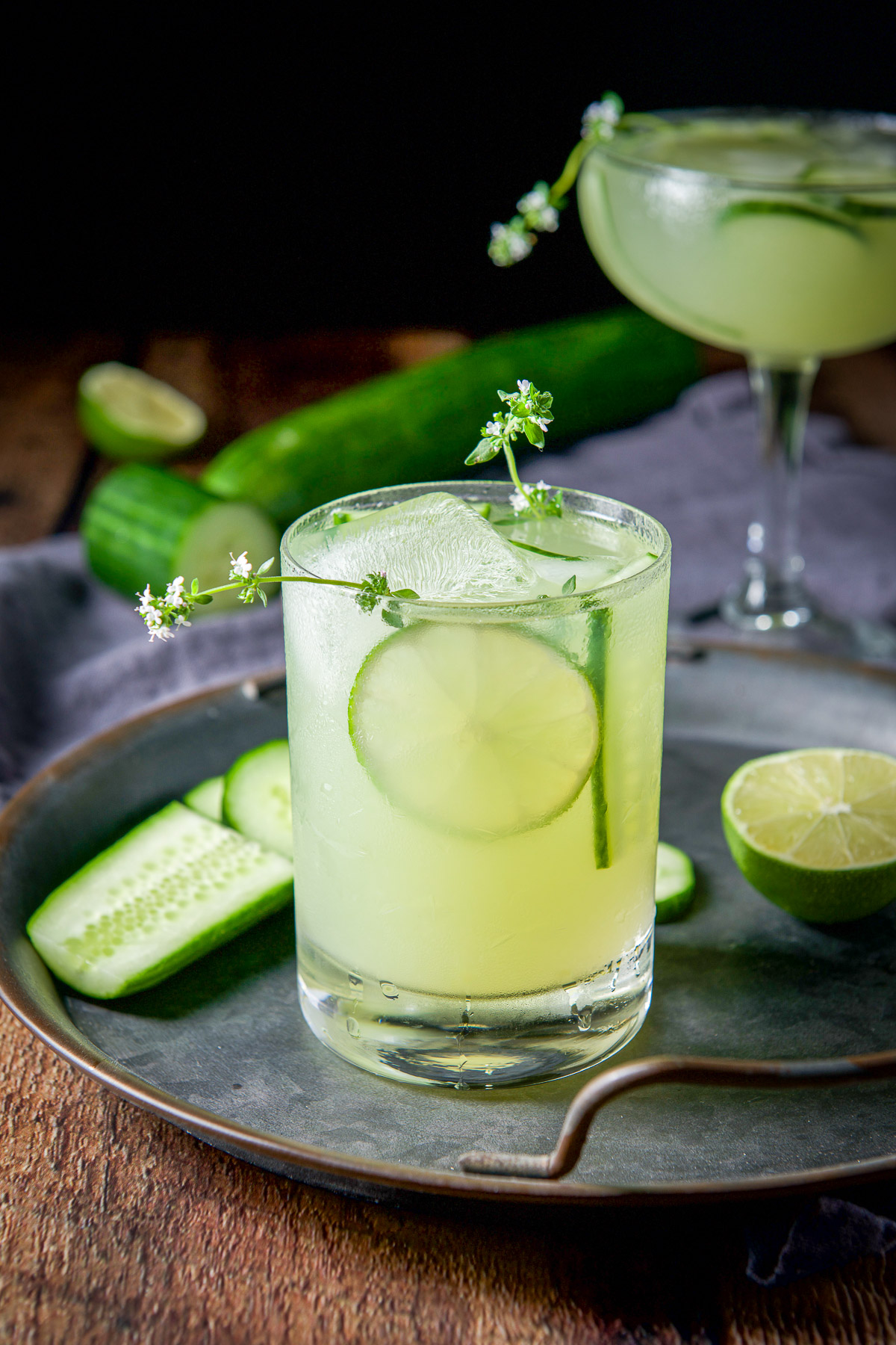 A glass on a tray filled with the margarita with fresh thyme, lime, and cucumber