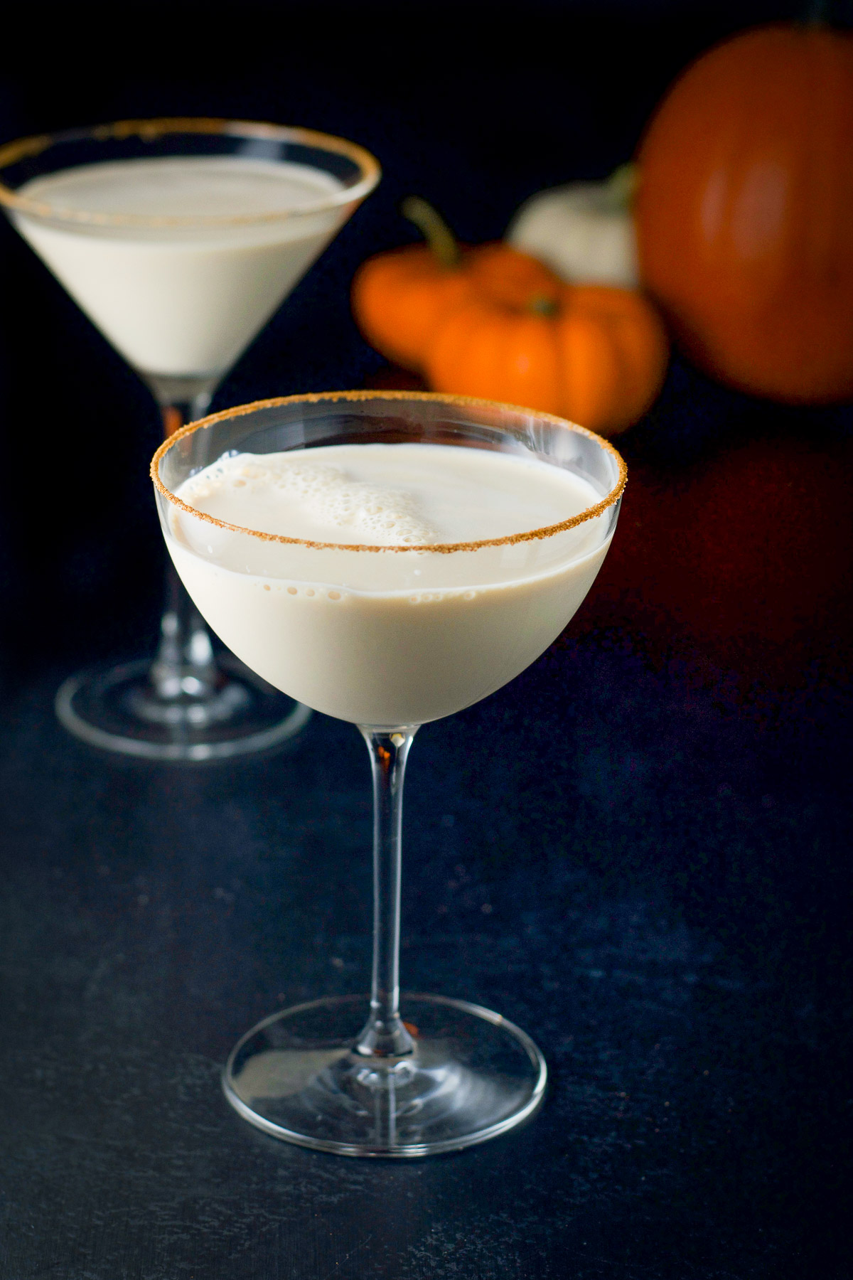 Two martini glasses filled with the cream martini with pumpkins in the back