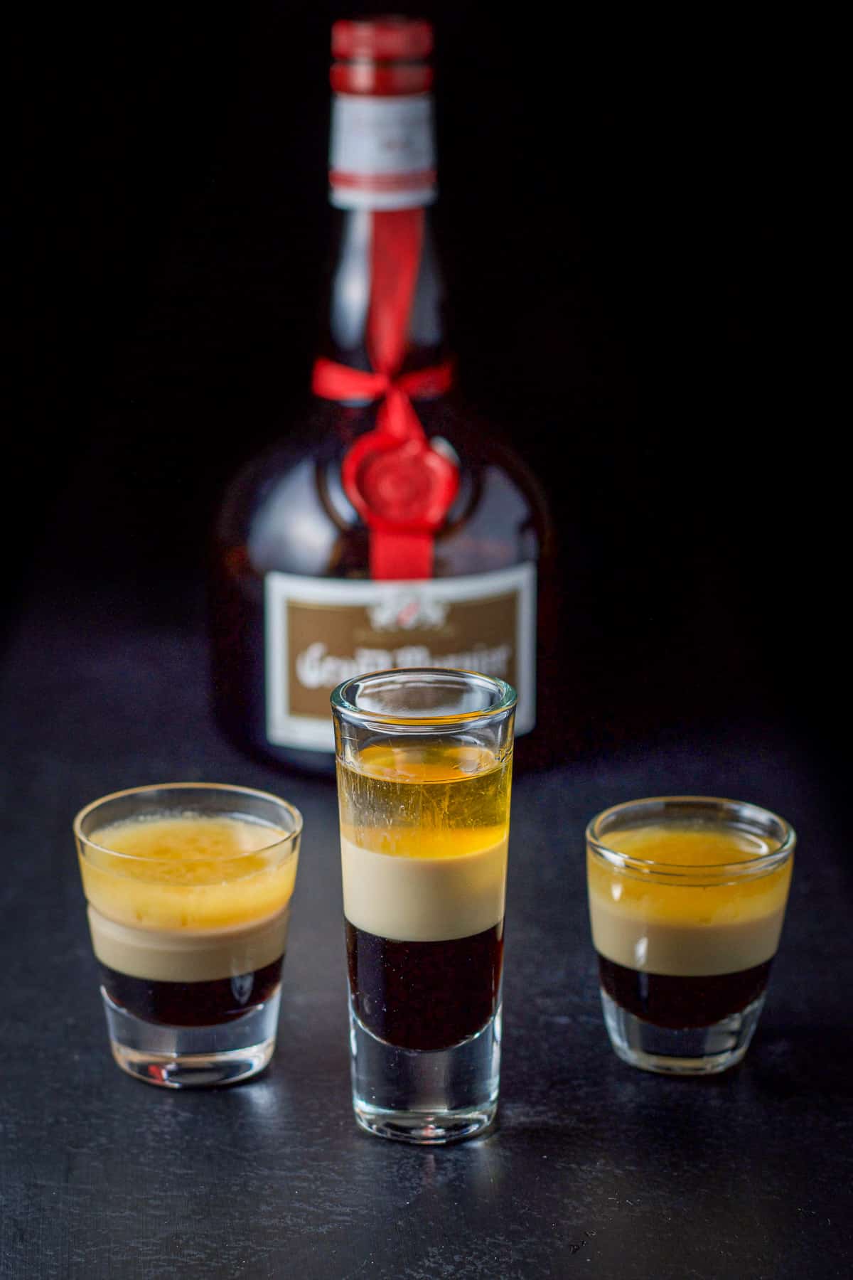 3 glasses filled with a layered shot with a bottle in the back