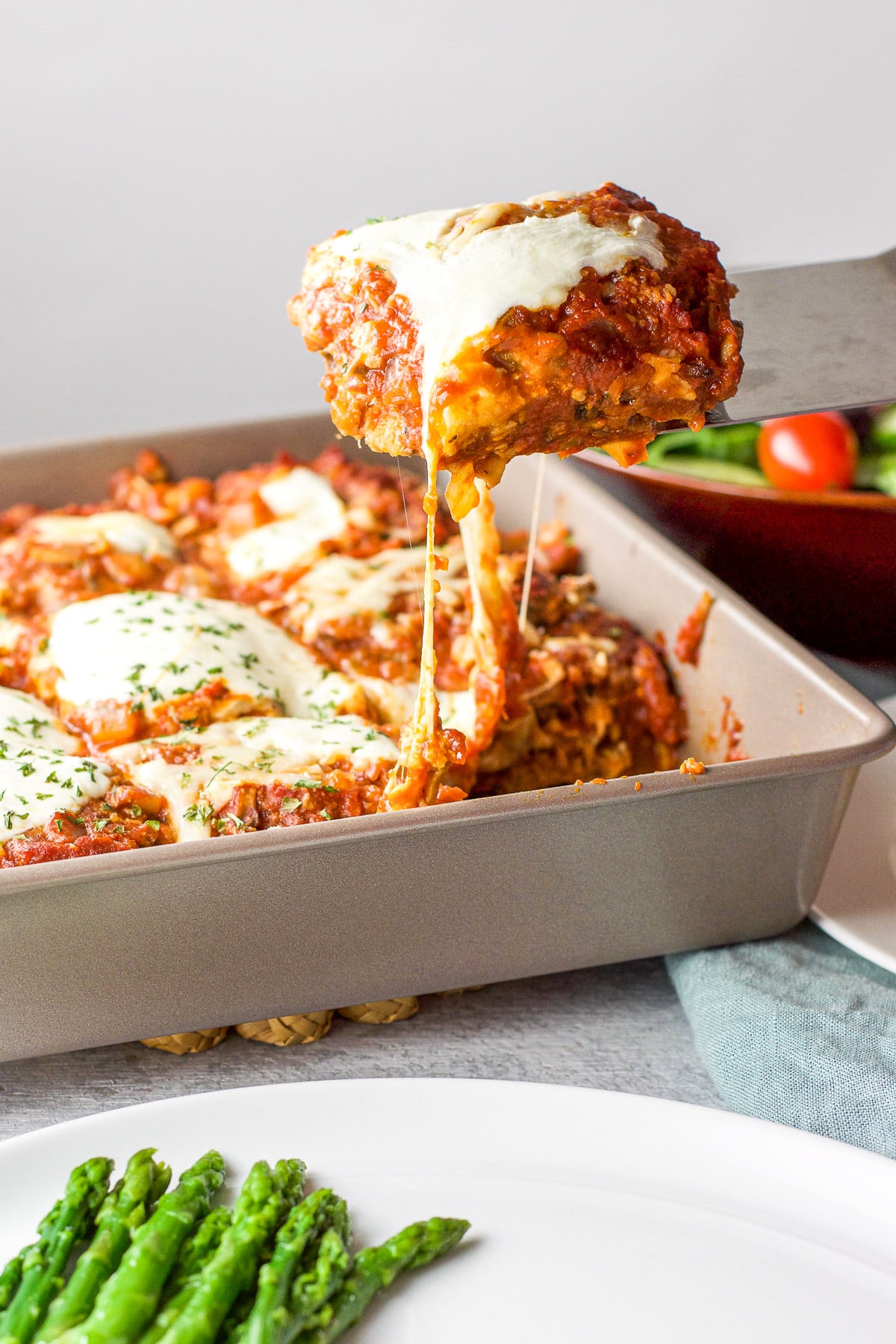 A spatula holding eggplant parm held over a plate with the pan in the back