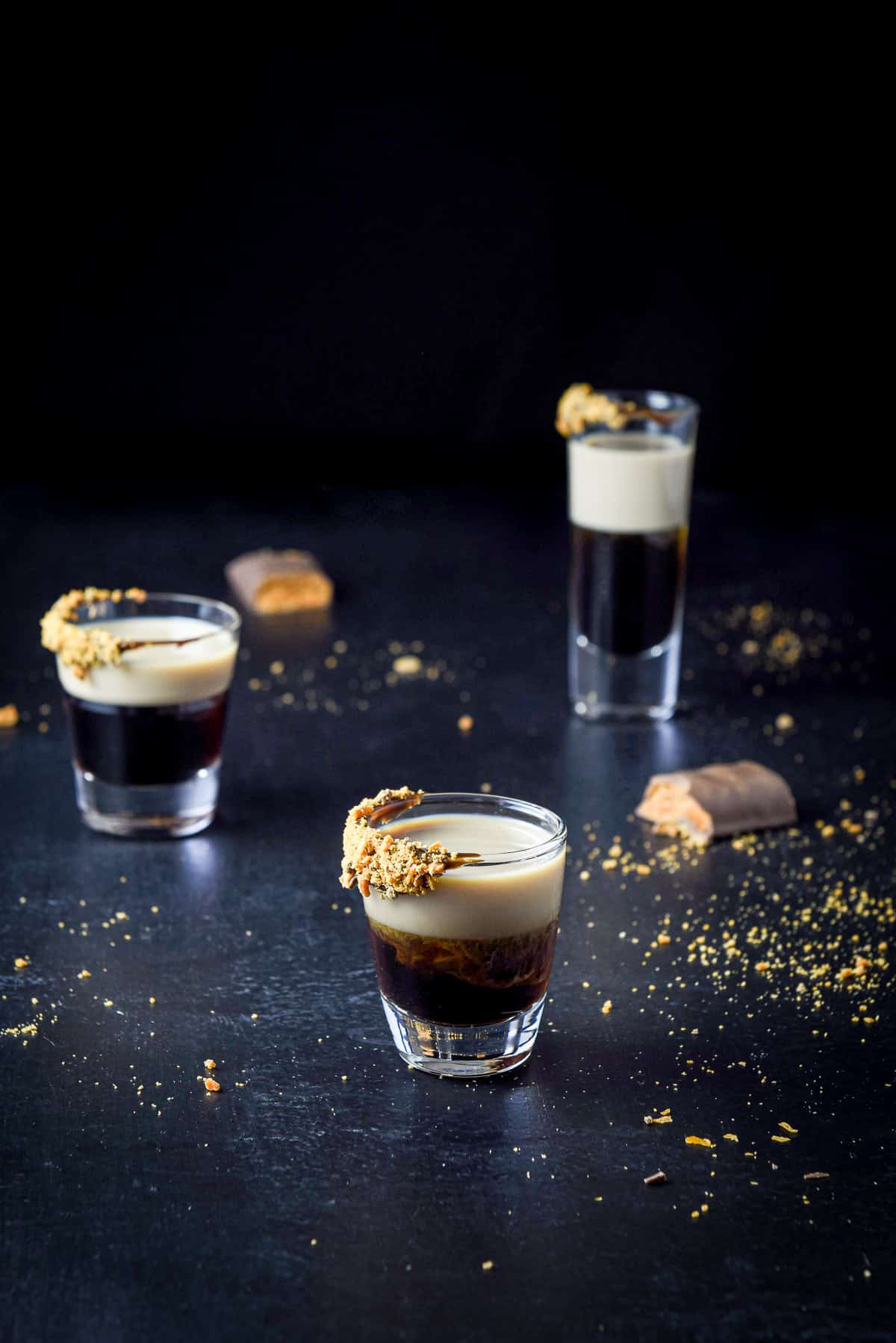Short glass in front of two other glasses with the shot it in with butterfinger bars on the table