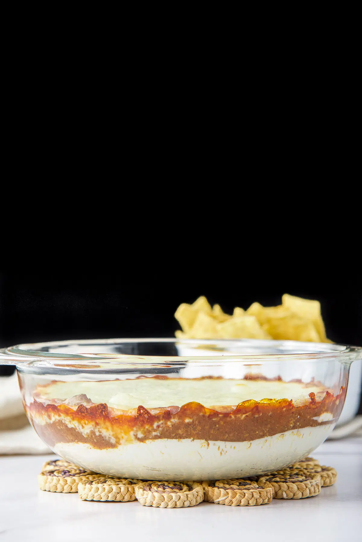 Vertical view of a casserole dish with the layered dip in it with chips in the back
