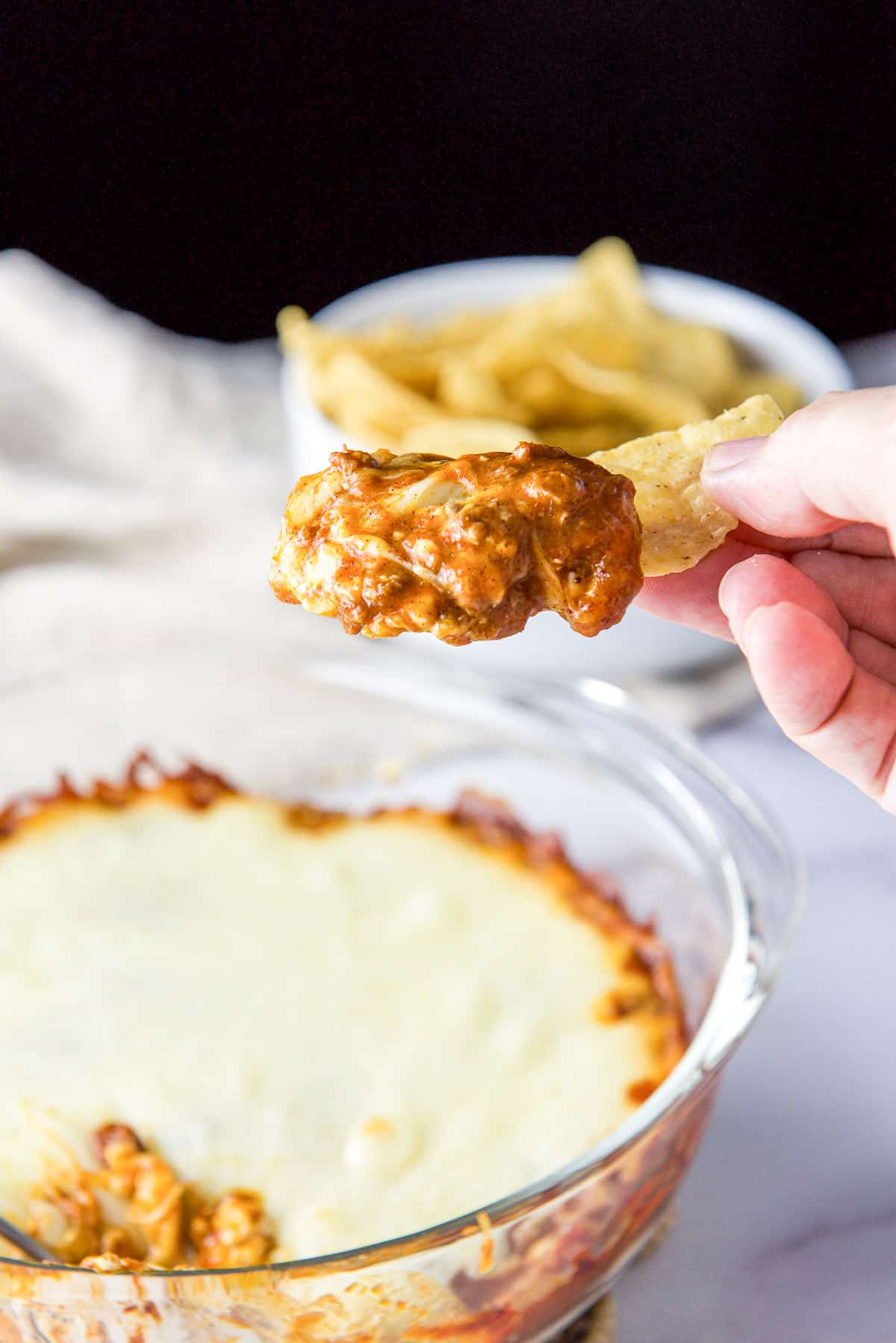A hand holding a chip with chili dip on it held over the casserole dish