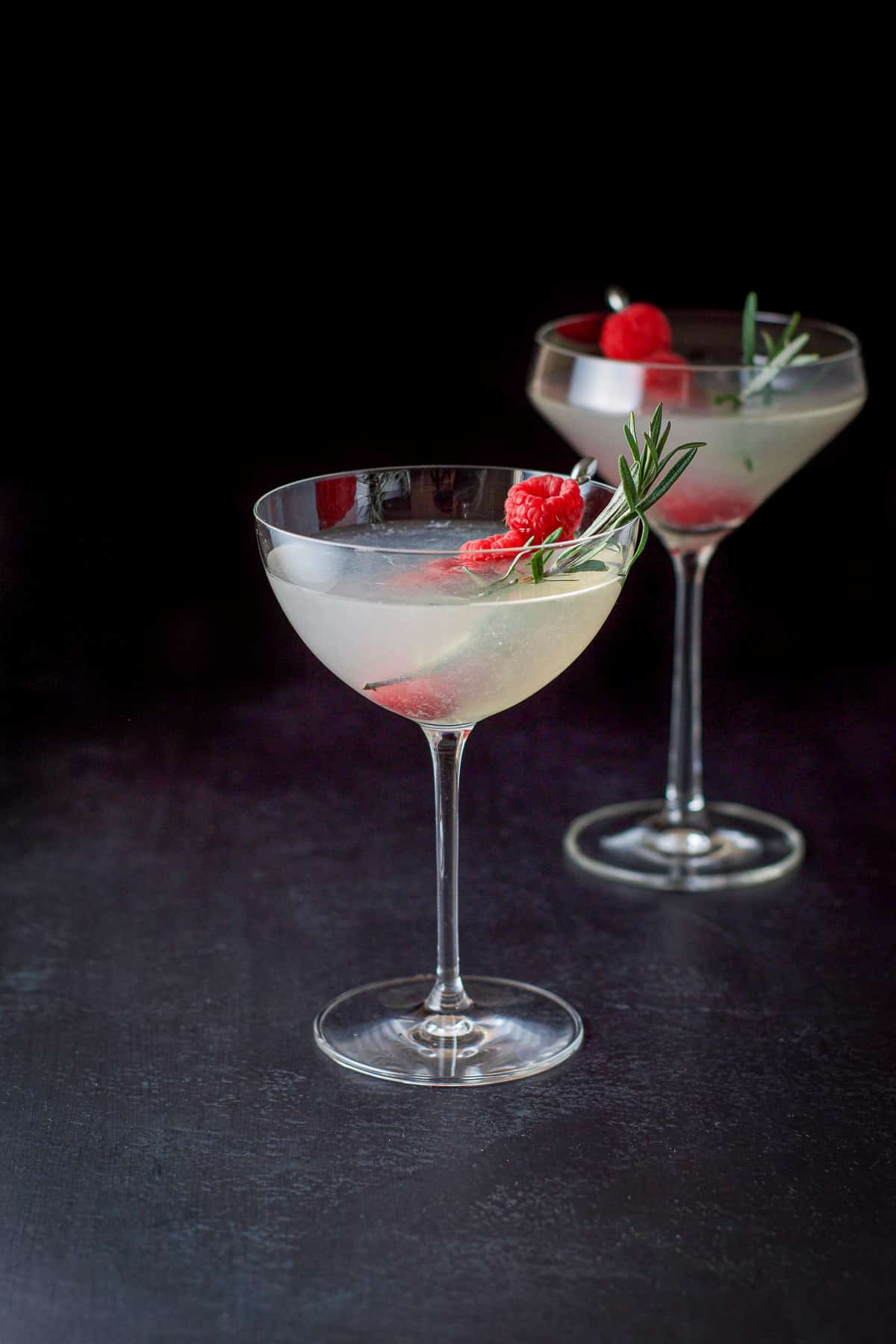 Two glasses filled with a mistletoe martini with raspberries and rosemary as garnish