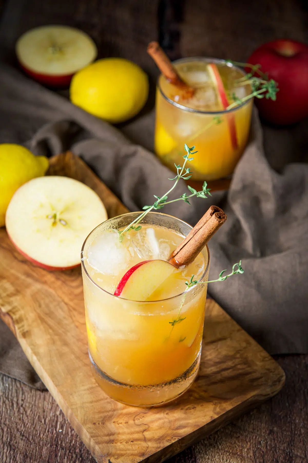 A glass on a wooden board filled with the apple bourbon smash with more apples and lemons