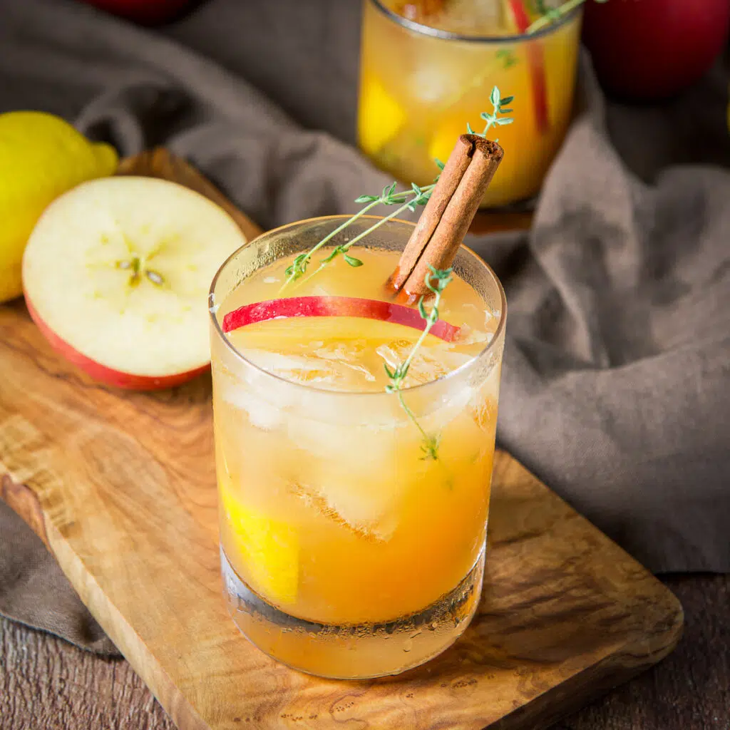 Square photo of a wood board with an apple cider drink in a glass