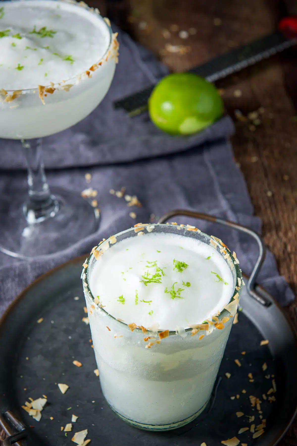 Two margarita glasses with the drinks in them with toasted coconut with lime shavings