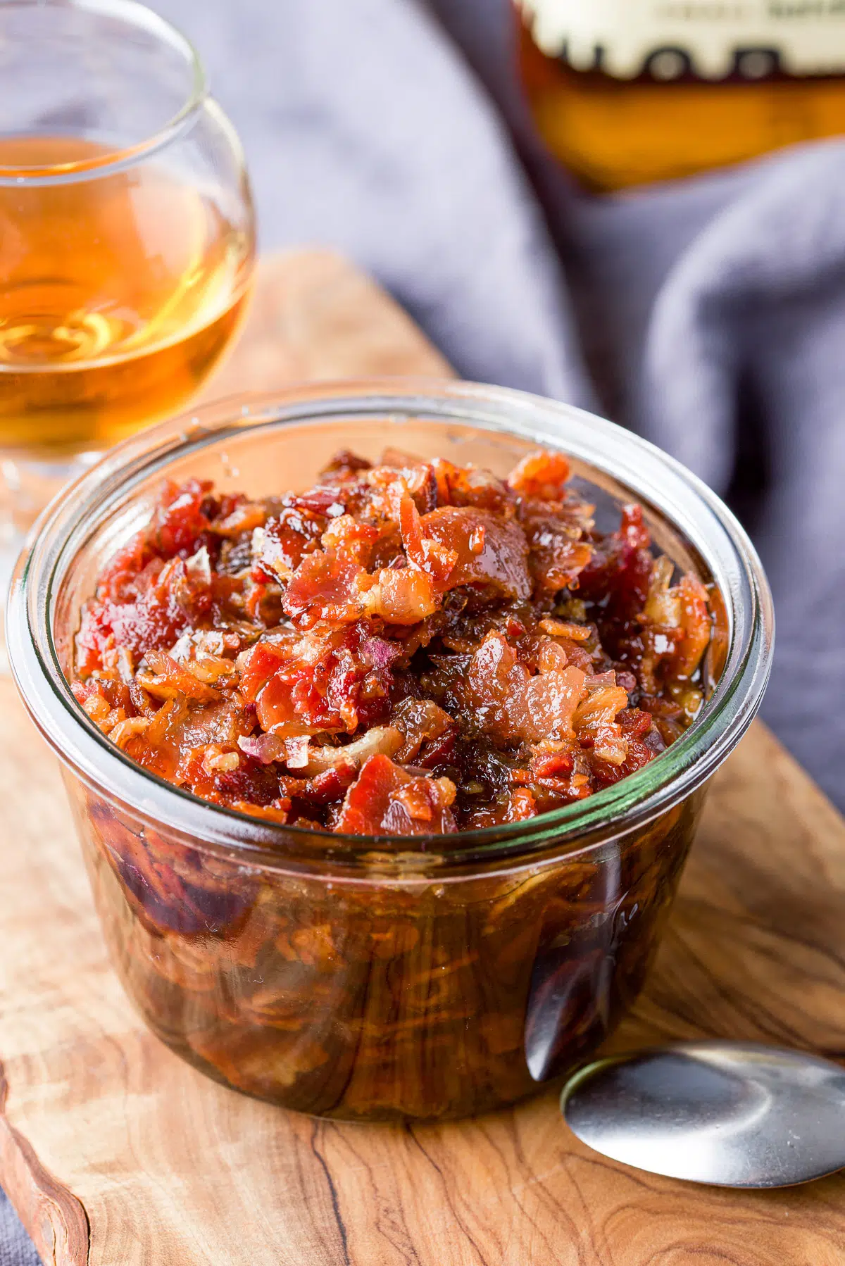 A wooden board with a spoon, bacon jam in a jar and bourbon in a glass
